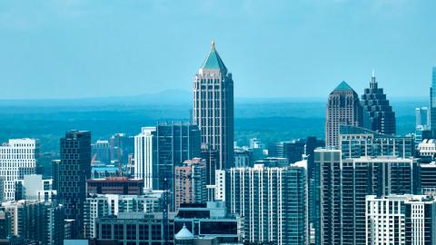 A skyline photo of Atlanta under blue skies with many tall buildings and highways and mountains in the far distance.