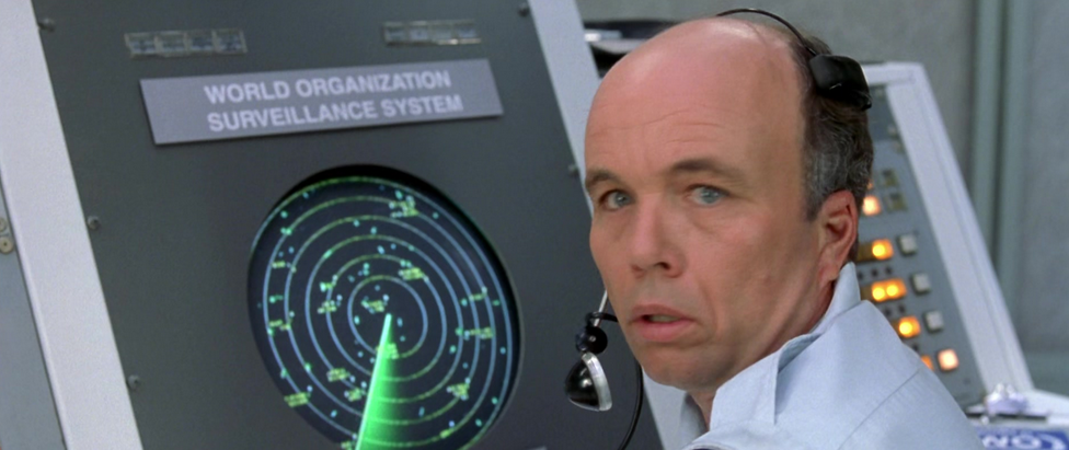 Screenshot of the 2002 film Austin Powers in Goldmember showing radar operator Johnson Ritter (played by Clint Howard) wearing a headset with a microphone. He has a concerned facial expression as he looks over his shoulder away from his circular radar display. Above the display is a placard with the text "WORLD ORGANIZATION SURVEILLANCE SYSTEM".