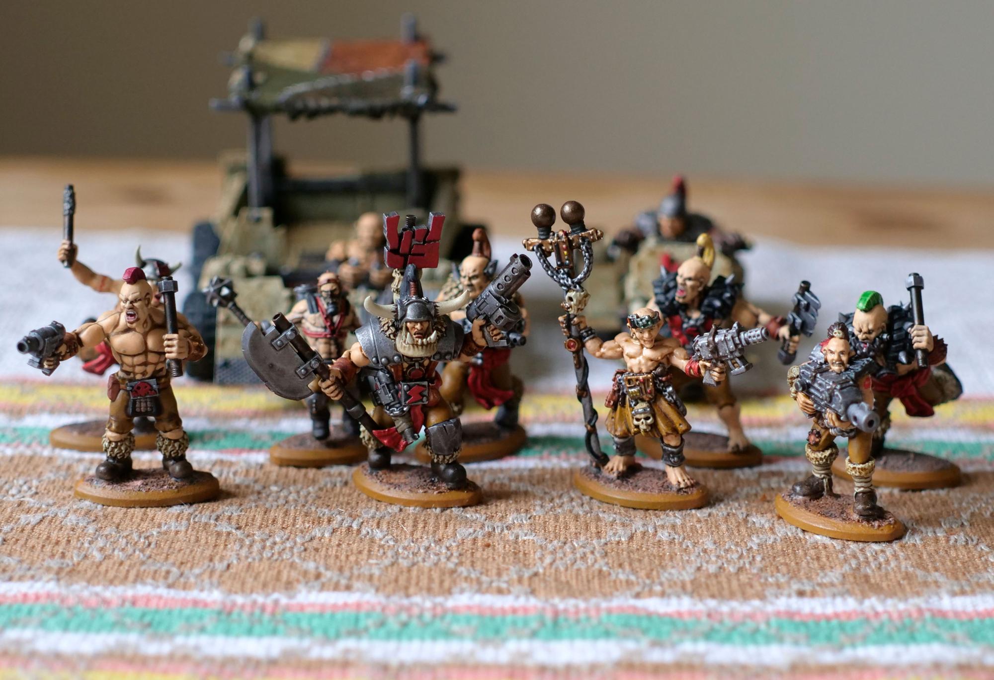 A group of miniatures wearing post-apocalyptic raider clobber. They all have tanned white skin and in the background are two similarly ramshackle vehicles.